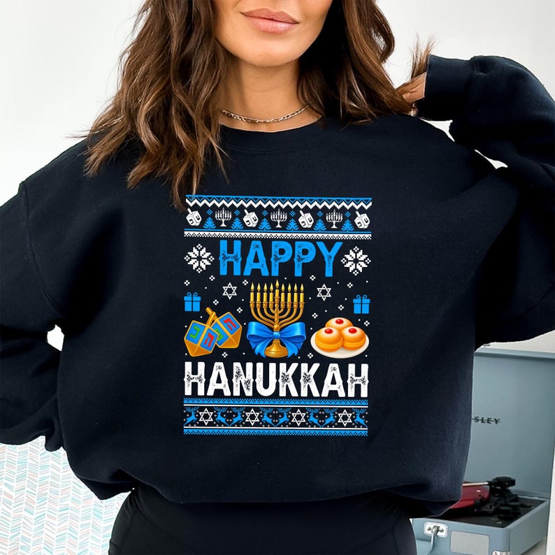 Może przedstawiać: Czarna bluza z świątecznym wzorem Chanuki. Wz&oacute;r zawiera słowa "HAPPY HANUKKAH" w kolorze niebieskim, wraz z menorami, bączkami, prezentami i sufganiyot. Projekt ma niebiesko-białą kolorystykę.
