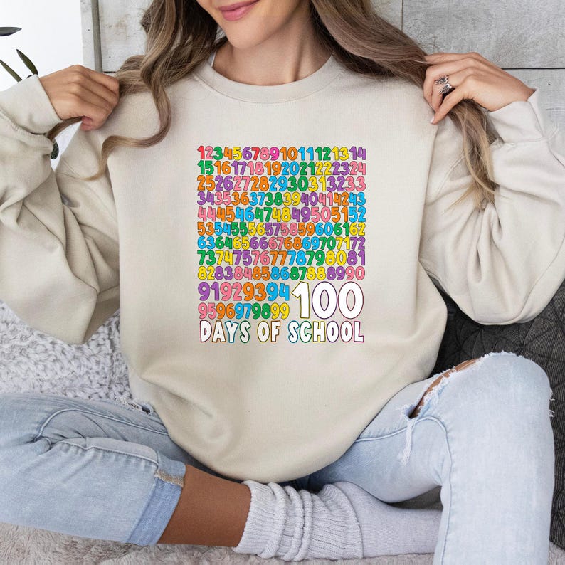 May include: A beige sweatshirt with a colorful graphic celebrating 100 days of school. The graphic includes numbers and the text "100 DAYS OF SCHOOL" in a rainbow of colors. The sweatshirt is a casual, comfortable style.
