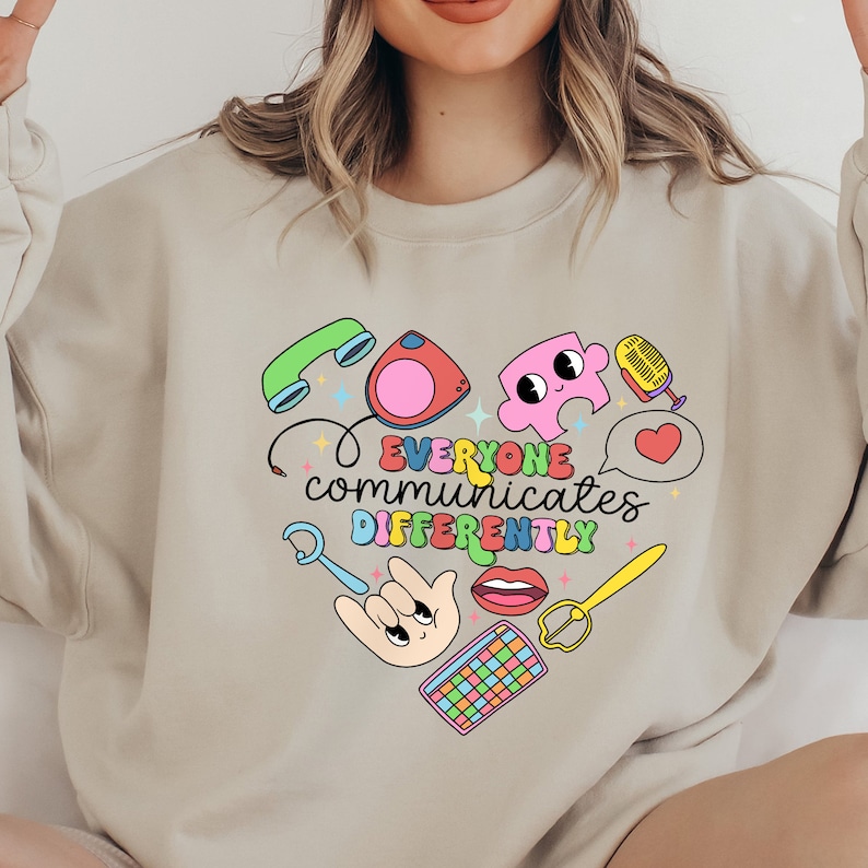 May include: A light beige sweatshirt with a graphic design of a heart shape made up of various colorful icons, including a phone, a microphone, a puzzle piece, a heart, a hand, a pair of lips, a pair of scissors, and a keyboard. The text "EVERYONE communicates DIFFERENTLY" is written in a colorful font inside the heart.
