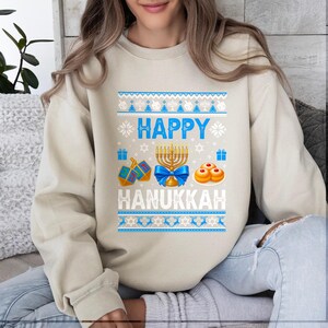 Może przedstawiać: Beżowa bluza z uroczym wzorem na Chanukę. Wz&oacute;r zawiera niebieskie słowa "Happy Hanukkah", a także menorę, bączki, prezenty i pączki. G&oacute;rna i dolna część wzoru mają niebieskie i białe wzory.