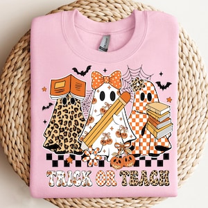 May include: Pink sweatshirt with a Halloween-themed graphic. The design features three ghost characters, one in leopard print, one with a bow and pencil, and one with a checkered pattern. The text "TRICK OR TEACH" is at the bottom.
