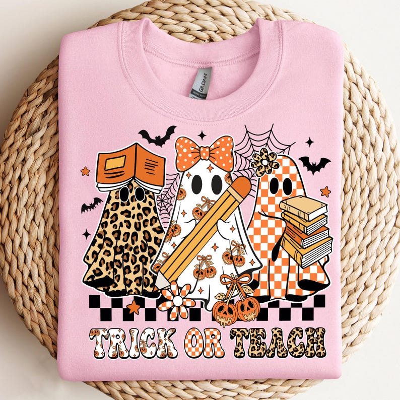 May include: Pink sweatshirt with a Halloween-themed graphic. The design features three ghost characters, one in leopard print, one with a bow and pencil, and one with a checkered pattern. The text "TRICK OR TEACH" is at the bottom.