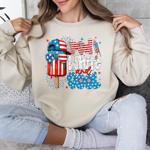 May include: A cream-colored sweatshirt featuring a patriotic popsicle design with sunglasses and a baseball cap. The text "Red, White, and Cool" is displayed in red, white, and blue colors. Fireworks and stars complete the design.