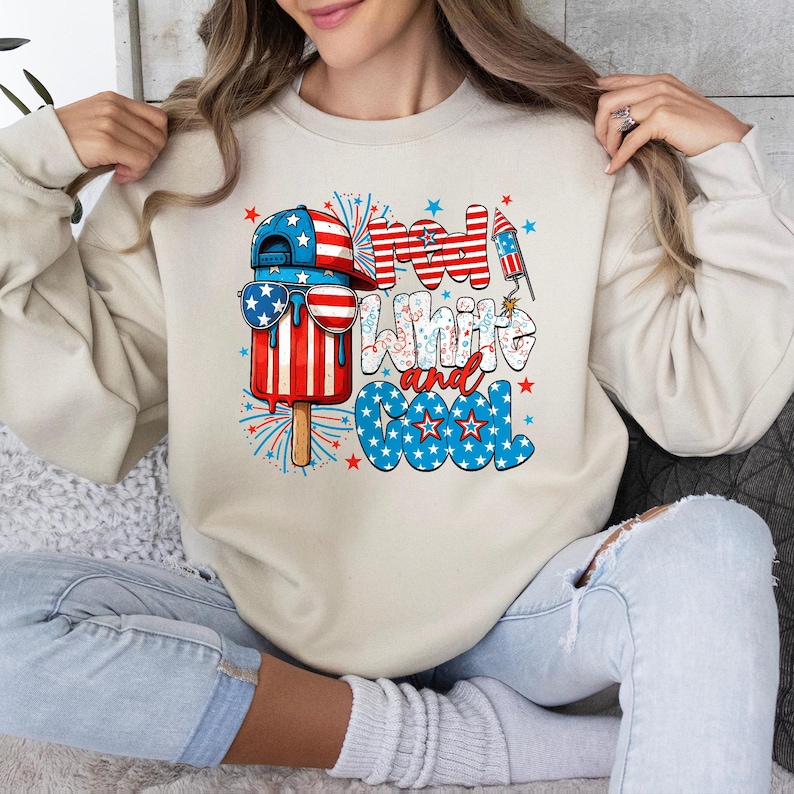 May include: A cream-colored sweatshirt featuring a patriotic popsicle design with sunglasses and a baseball cap. The text "Red, White, and Cool" is displayed in red, white, and blue colors. Fireworks and stars complete the design.