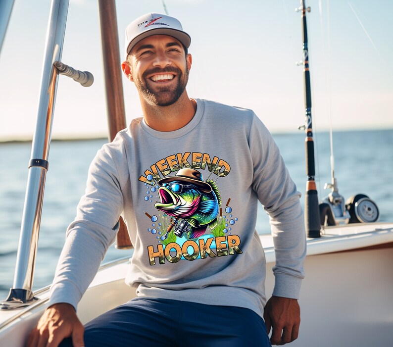 K&ouml;nnte beinhalten: Ein graues Sweatshirt mit einem bunten Cartoon-Fisch, der eine Sonnenbrille und einen Hut tr&auml;gt. Der Text "Weekend Hooker" ist auf dem Shirt aufgedruckt.