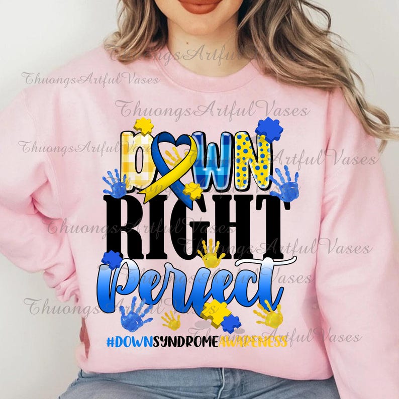 May include: Pink sweatshirt with a graphic design that says "Down Right Perfect" in black letters with a yellow and blue ribbon, blue puzzle pieces, and blue handprints. The text "#DownSyndromeAwareness" is at the bottom of the design.