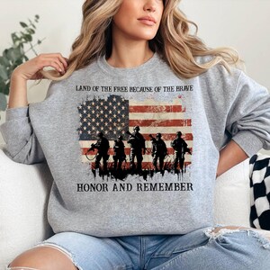 May include: A gray sweatshirt featuring an American flag design with the text "LAND OF THE FREE BECAUSE OF THE BRAVE" and "HONOR AND REMEMBER." The flag design includes silhouettes of soldiers. The sweatshirt is a comfortable, casual style.