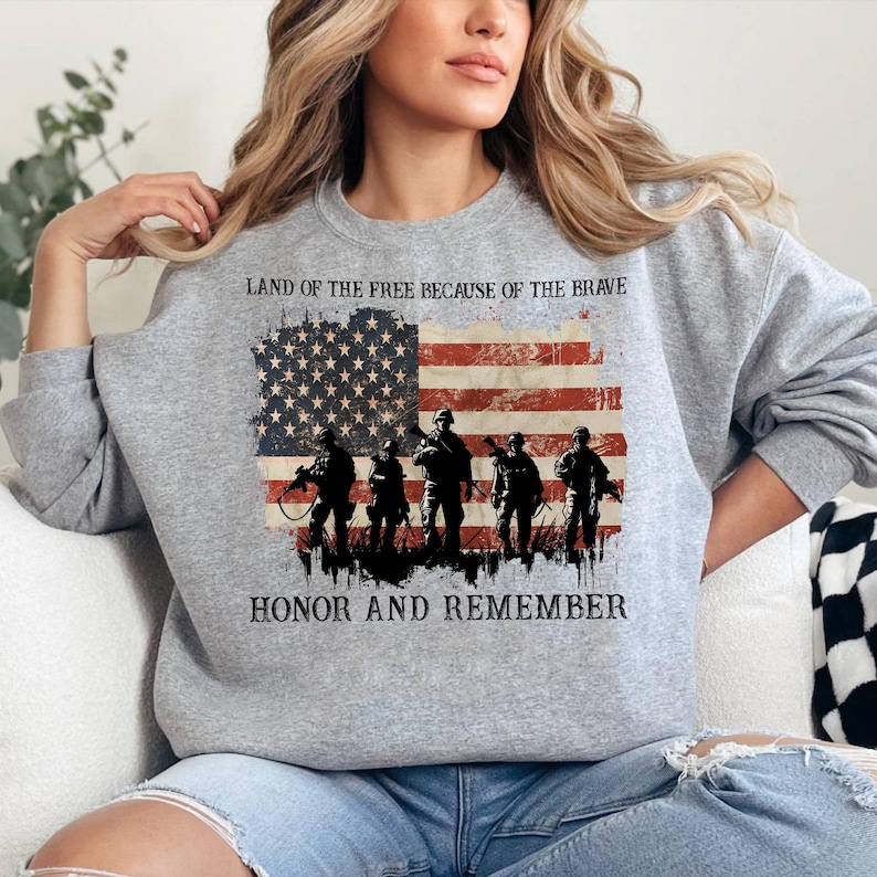 May include: A gray sweatshirt featuring an American flag design with the text "LAND OF THE FREE BECAUSE OF THE BRAVE" and "HONOR AND REMEMBER." The flag design includes silhouettes of soldiers. The sweatshirt is a comfortable, casual style.