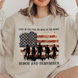 May include: A beige sweatshirt featuring a distressed American flag design with soldiers silhouetted in front. The text "LAND OF THE FREE BECAUSE OF THE BRAVE" and "HONOR AND REMEMBER" are printed above and below the flag.