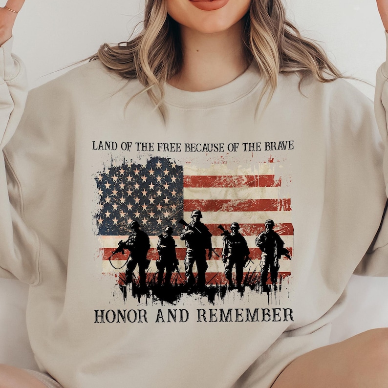 May include: A beige sweatshirt featuring a distressed American flag design with soldiers silhouetted in front. The text "LAND OF THE FREE BECAUSE OF THE BRAVE" and "HONOR AND REMEMBER" are printed above and below the flag.