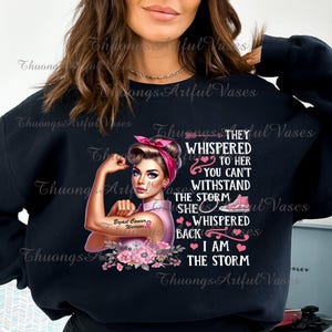 Puede incluir: Una sudadera negra con un dise&ntilde;o gr&aacute;fico rosa y blanco que representa a una mujer con una pose de brazo fuerte. El texto dice "Le susurraron a ella que no puedes resistir la tormenta, ella susurr&oacute; de vuelta, yo soy la tormenta". El gr&aacute;fico tambi&eacute;n incluye una cinta rosa con las palabras "Guerrera del c&aacute;ncer de mama".