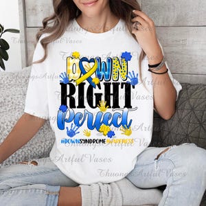 May include: White t-shirt with the text "Down Right Perfect" in black and blue lettering. The text is surrounded by yellow and blue handprints. The shirt also includes a yellow and blue ribbon with a heart in the center. The text "#DownSyndromeAwareness" is printed below the main text.