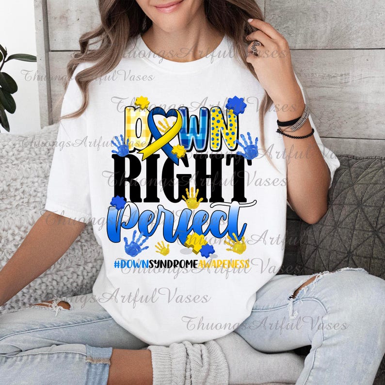 May include: White t-shirt with the text "Down Right Perfect" in black and blue lettering. The text is surrounded by yellow and blue handprints. The shirt also includes a yellow and blue ribbon with a heart in the center. The text "#DownSyndromeAwareness" is printed below the main text.