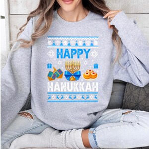 Może przedstawiać: Jasnoszara bluza z świątecznym wzorem Chanuki. Projekt zawiera słowa "HAPPY HANUKKAH" w kolorze białym i niebieskim, a także obrazy menory, bączk&oacute;w i sufganiyot. G&oacute;rna i dolna część projektu mają niebiesko-biały wz&oacute;r.