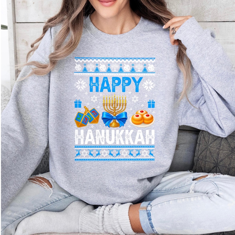 Może przedstawiać: Jasnoszara bluza z świątecznym wzorem Chanuki. Projekt zawiera słowa "HAPPY HANUKKAH" w kolorze białym i niebieskim, a także obrazy menory, bączk&oacute;w i sufganiyot. G&oacute;rna i dolna część projektu mają niebiesko-biały wz&oacute;r.