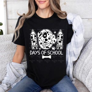 May include: Black and white graphic design of a Dalmatian dog with a tongue sticking out. The dog is surrounded by two pencils and the text "100 Days of School" with a bone underneath.