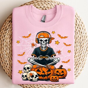 May include: Pale pink sweatshirt with a skeleton gamer design. The skeleton wears headphones and an orange cap, holding a controller. The design includes pumpkins, bats, spiders, stars, and spiderwebs, ideal for Halloween.