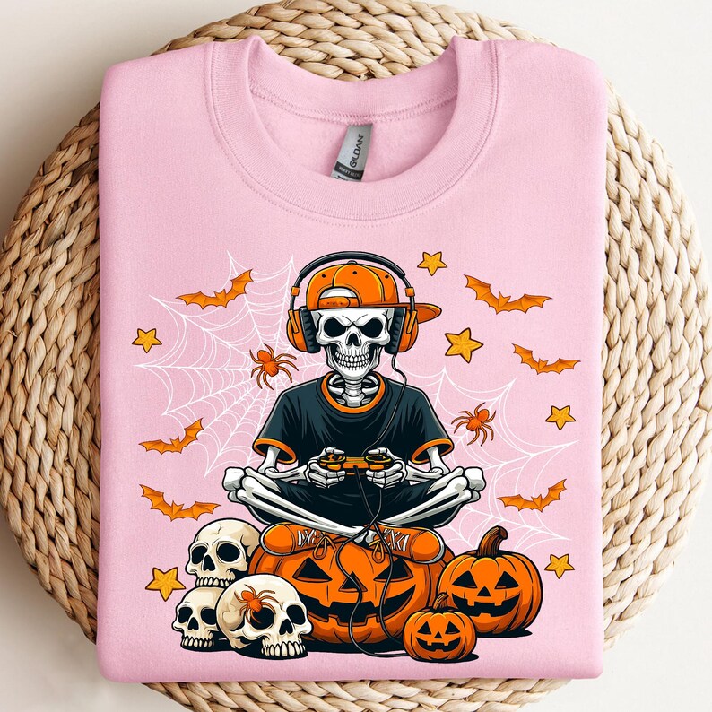 May include: Pale pink sweatshirt with a skeleton gamer design. The skeleton wears headphones and an orange cap, holding a controller. The design includes pumpkins, bats, spiders, stars, and spiderwebs, ideal for Halloween.