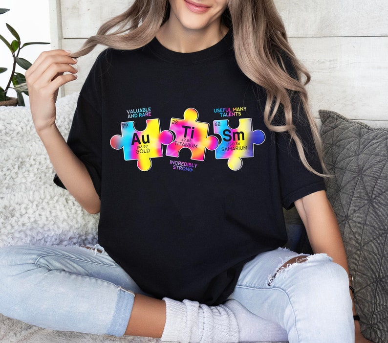 May include: A black t-shirt with a colourful graphic of jigsaw pieces featuring the chemical symbols for gold, titanium, and samarium. The text on the graphic reads "Valuable and Rare", "Incredibly Strong", and "Useful Many Talents".