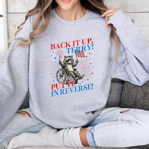 May include: A light gray sweatshirt featuring a raccoon in a wheelchair holding an American flag. The graphic includes the text "BACK IT UP, TERRY!" and "PUT IT IN REVERSE!" in red and blue, with fireworks.