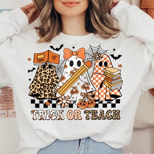 May include: White sweatshirt featuring three ghost characters with Halloween-themed designs. One ghost wears a leopard print, another holds a pencil, and the third holds books. The phrase "TRICK OR TEACH" is printed below the ghosts.
