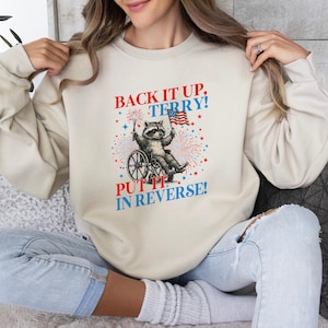 May include: Cream-colored sweatshirt with a graphic featuring a raccoon in a wheelchair holding an American flag and fireworks. Text reads "BACK IT UP, TERRY! PUT IT IN REVERSE!" in red and blue.