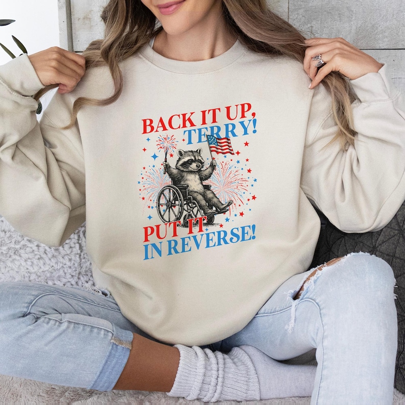 May include: Cream-colored sweatshirt with a graphic featuring a raccoon in a wheelchair holding an American flag and fireworks. Text reads "BACK IT UP, TERRY! PUT IT IN REVERSE!" in red and blue.