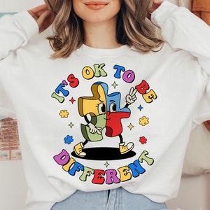 May include: White sweatshirt with a colorful graphic of a puzzle piece with a face and the text "It's OK to be different".
