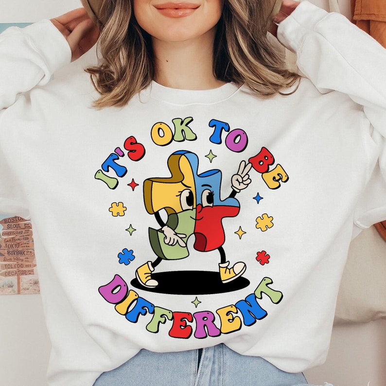 May include: White sweatshirt with a colorful graphic of a puzzle piece with a face and the text "It's OK to be different".