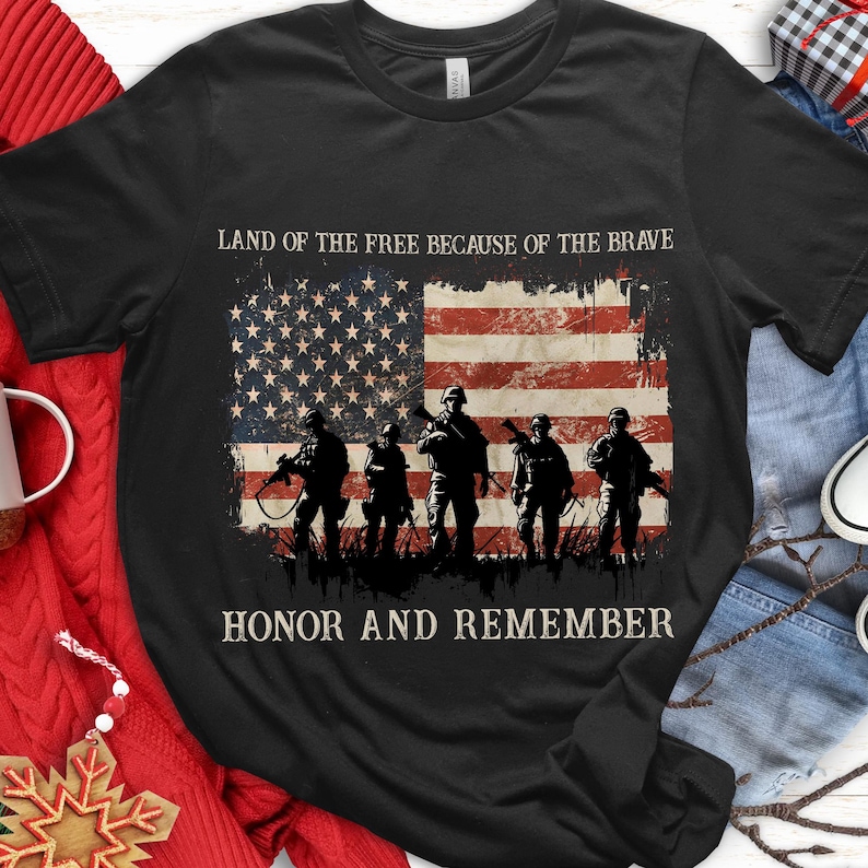 May include: Black t-shirt featuring an American flag design with soldiers in silhouette. The shirt includes the phrases "LAND OF THE FREE BECAUSE OF THE BRAVE" and "HONOR AND REMEMBER" in white text. The design is distressed.