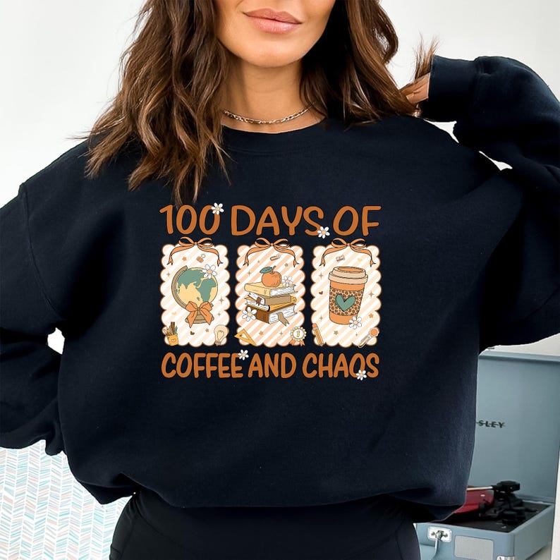 May include: Black sweatshirt with the text "100 Days of Coffee and Chaos." The design includes three framed illustrations: a globe, books with a pumpkin, and a coffee cup with a heart. The illustrations are in shades of brown, orange, and white.