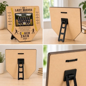 May include: A wooden baseball home plate-shaped plaque with a team photo and text. The plaque features the text "8U Lady Braves Softball" and "Coach Zach - 2026". The plaque has a black stand on the back.