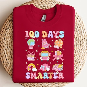 May include: A maroon sweatshirt with a colorful graphic celebrating 100 days of school. The design includes a globe, backpack, apple, school bus, and the word "SMARTER". The sweatshirt is made of a soft material.