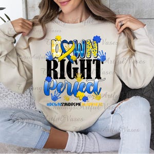 May include: A beige sweatshirt with the text "Down Right Perfect" in blue and yellow lettering. The text is surrounded by handprints in blue and yellow. The text "#DownSyndromeAwareness" is printed in smaller letters below the main text.