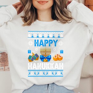 Może przedstawiać: Biała bluza z długim rękawem z niebiesko-białym wzorem w tematyce Chanuki. Projekt zawiera słowa "HAPPY HANUKKAH", menorę, dreidels i sufganiyot. Bluza ma okrągły dekolt.