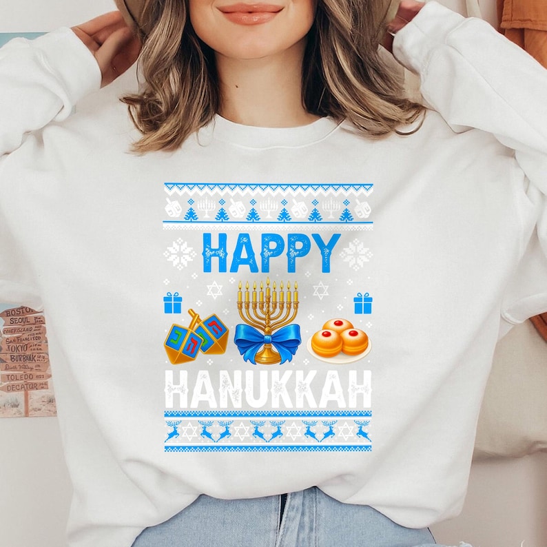 Może przedstawiać: Biała bluza z długim rękawem z niebiesko-białym wzorem w tematyce Chanuki. Projekt zawiera słowa "HAPPY HANUKKAH", menorę, dreidels i sufganiyot. Bluza ma okrągły dekolt.
