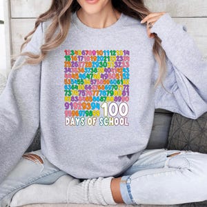 May include: A light gray sweatshirt with a colorful graphic celebrating 100 days of school. The design features a grid of numbers in various colors, with "100" and "Days of School" in white text below.