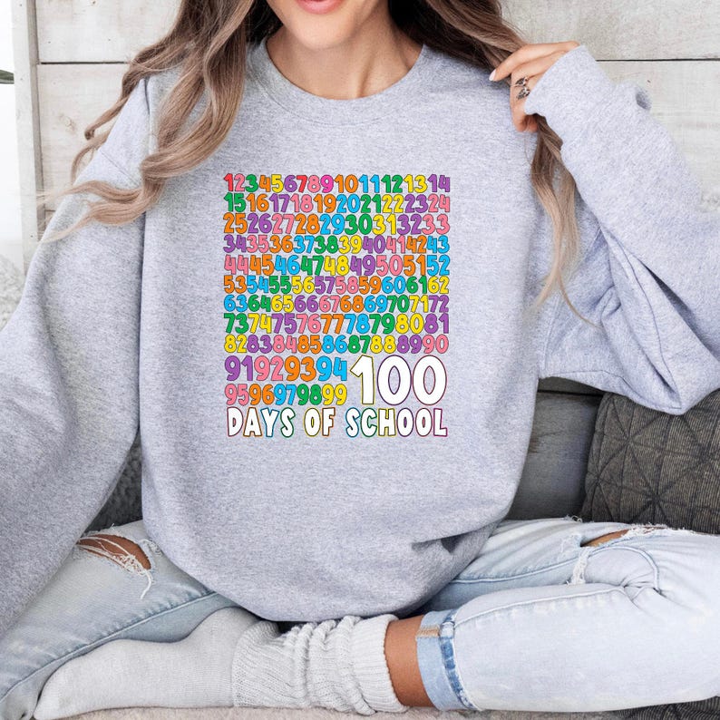 May include: A light gray sweatshirt with a colorful graphic celebrating 100 days of school. The design features a grid of numbers in various colors, with "100" and "Days of School" in white text below.
