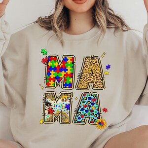 May include: A beige sweatshirt with the word "MAMA" written in large letters. Each letter is made up of different patterns, including jigsaw pieces, leopard print, and hearts.