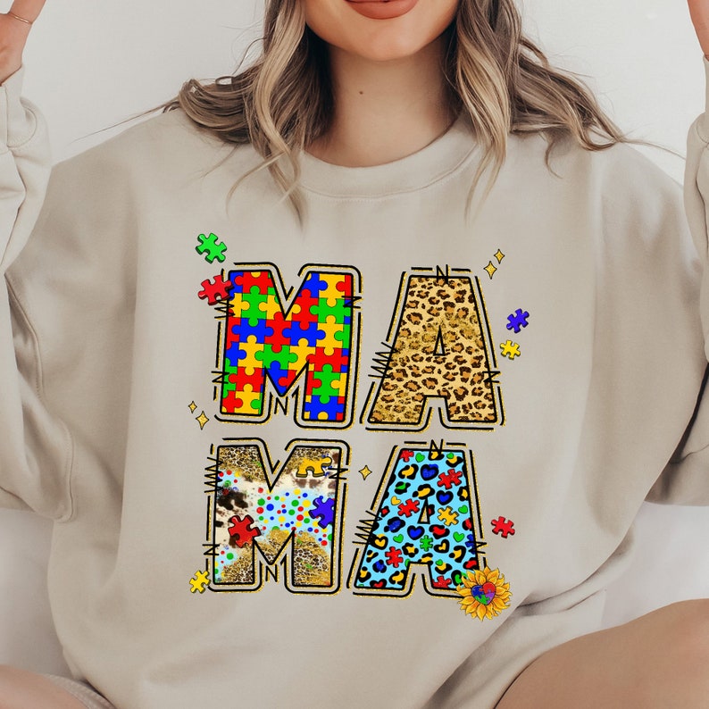 May include: A beige sweatshirt with the word "MAMA" written in large letters. Each letter is made up of different patterns, including jigsaw pieces, leopard print, and hearts.