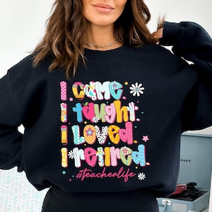 May include: Black sweatshirt with the text "I came, I taught, I loved, I retired" in colorful, patterned letters. The phrase "#teacherlife" is below the text, with stars and flower accents. A casual, comfortable style.