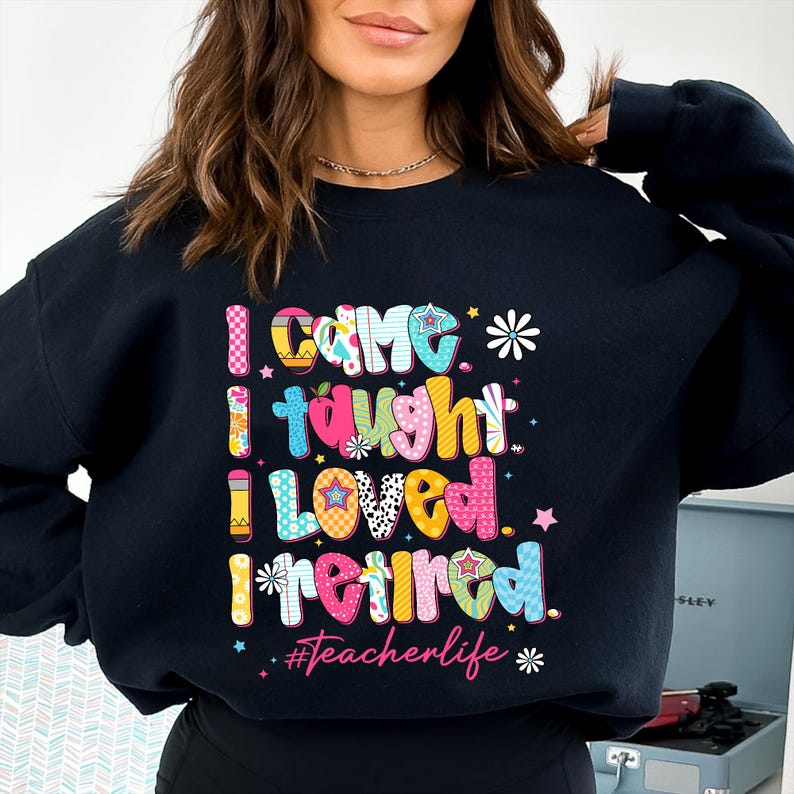 May include: Black sweatshirt with the text "I came, I taught, I loved, I retired" in colorful, patterned letters. The phrase "#teacherlife" is below the text, with stars and flower accents. A casual, comfortable style.