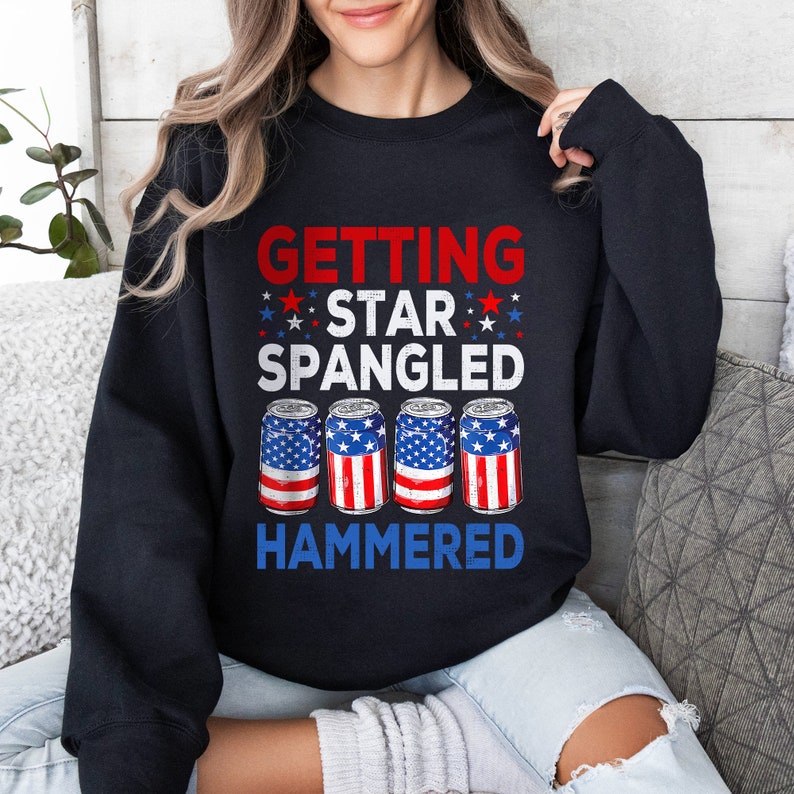 May include: Black sweatshirt with a red, white, and blue American flag design. The text "GETTING STAR SPANGLED HAMMERED" is printed on the shirt. Four cans of beer with American flag designs are printed below the text.