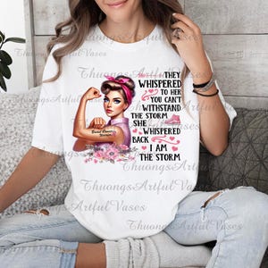 Puede incluir: Camiseta blanca con un gr&aacute;fico rosa y blanco de una mujer con un brazo fuerte, una cinta rosa y el texto "They whispered to her you can't withstand the storm she whispered back I am the storm".