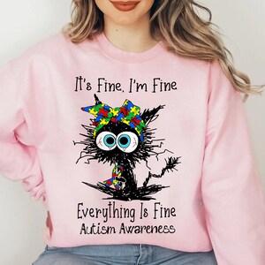 May include: Pink sweatshirt with a black cat illustration wearing a headband with puzzle pieces in the colors of the autism awareness ribbon. The text "It's Fine. I'm Fine. Everything Is Fine. Autism Awareness" is printed on the sweatshirt.