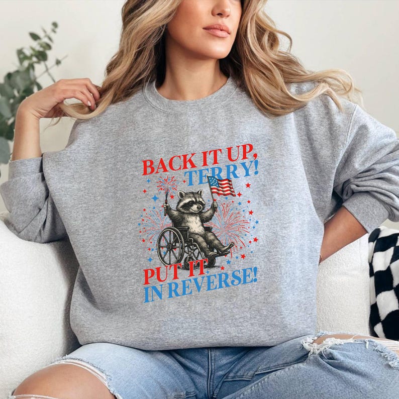 May include: A gray sweatshirt featuring a raccoon in a wheelchair holding a US flag and fireworks. The text "BACK IT UP, TERRY! PUT IT IN REVERSE!" is printed in red and blue. The design includes red and blue stars.