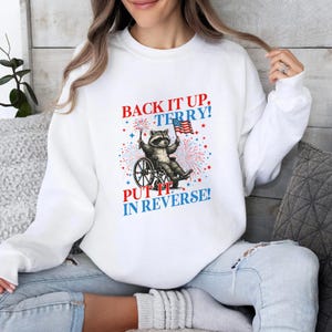 May include: White sweatshirt featuring a raccoon in a wheelchair holding an American flag and a sparkler. The text "BACK IT UP, TERRY! PUT IT IN REVERSE!" is printed in red and blue. The design includes fireworks.
