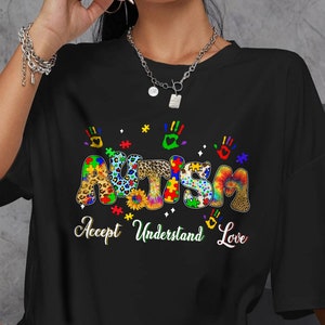 May include: Black t-shirt featuring a colourful graphic of the word "Autism" made up of jigsaw pieces, animal print, and tie-dye patterns. The text "Accept Understand Love" is below the graphic.