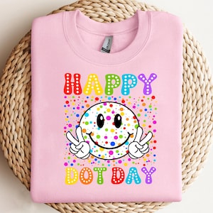 May include: A light pink sweatshirt with a colorful graphic. The graphic features the words "HAPPY DOT DAY" in rainbow colors, a smiley face with peace signs, and a background of colorful dots.
