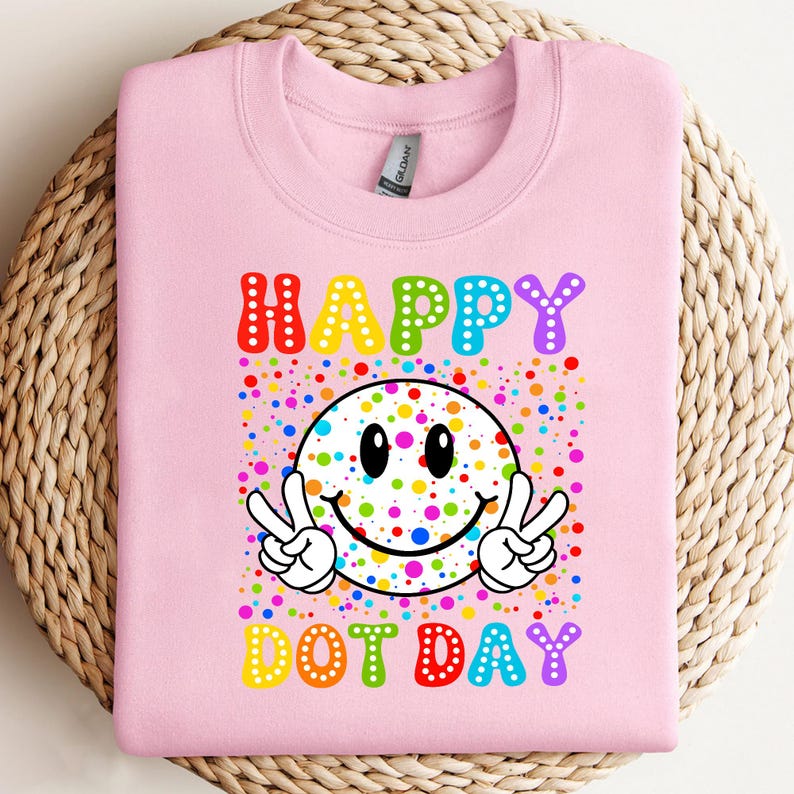 May include: A light pink sweatshirt with a colorful graphic. The graphic features the words "HAPPY DOT DAY" in rainbow colors, a smiley face with peace signs, and a background of colorful dots.
